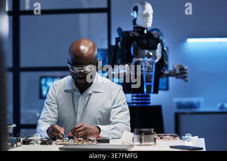 Ingegnere informatico maschio nero in camice da laboratorio e occhiali protettivi che lavorano sulla scheda madre seduto al tavolo di fronte al robot in laboratorio Foto Stock