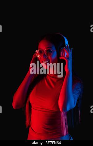 Donna asiatica con occhiali da sole a forma di cuore che ascolta musica con le cuffie in piedi su sfondo nero con luce rossa in studio Foto Stock