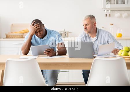 Uomo nero stressato che si appoggia alla testa e usa il tablet seduto a tavola con un amico di mezza età caucasico che lavora su documenti e grembo... Foto Stock