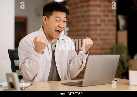Allegro uomo d'affari asiatico che guarda lo schermo del notebook e trema i pugni stringuti che celebrano le buone notizie seduto al tavolo di legno a casa Foto Stock