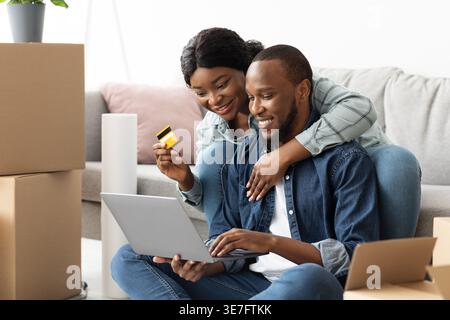 Moglie nera che tiene in mano la carta di credito e guarda il computer portatile che utilizza il marito, mentre è seduta insieme sul pavimento nel soggiorno vicino alle scatole non imballate di casa Foto Stock