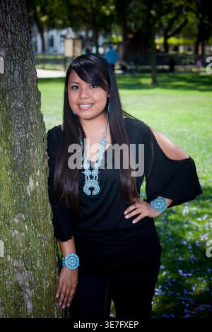 Donna sorridente dei nativi americani che indossa gioielli turchesi in piedi con la mano sulla vita appoggiata contro l'albero nel parco durante la giornata di sole Foto Stock