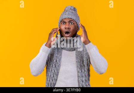 Scioccato uomo Nero che indossa berretto e sciarpa a maglia e che riceve una telefonata guardando lontano in piedi sullo sfondo giallo in studio Foto Stock