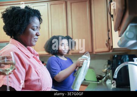 La figlia nera con afro che usa un asciugamano da cucina nelle piastre di asciugatura aiutando sua madre mentre si trova vicino al lavandino della cucina a casa Foto Stock