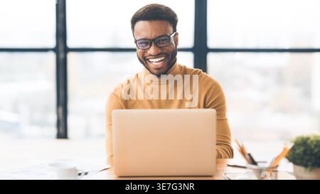 Allegro uomo nero che indossa occhiali da vista seduto a un tavolo con un computer portatile in ufficio Foto Stock