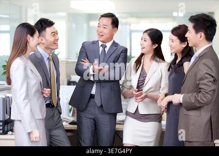 Uomo d'affari cinese di mezza età sorridente con le braccia incrociate in piedi e che parla con colleghi di sesso maschile e femminile in carica Foto Stock