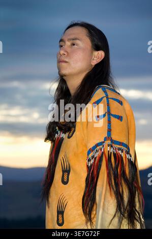 Serio uomo nativo americano con capelli lunghi e camicia in buckskin con perline che guarda lontano in piedi sulla montagna al tramonto Foto Stock