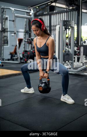 La donna thailandese in forma fisica si veste ascoltando musica con le cuffie e guardando in basso mentre esegue squat kettlebell in palestra Foto Stock