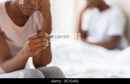 Donna nera appoggiata faccia a mano tenendo il kit test di gravidanza seduta sul letto vicino al marito in camera da letto a casa Foto Stock
