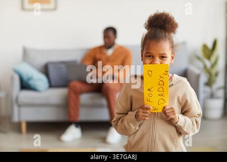 Una ragazza nera che regge un regalo a sorpresa fatto a mano per suo padre mentre si trova in salotto a casa Foto Stock