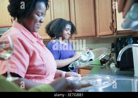 La figlia nera con afro che usa un asciugamano da cucina nelle piastre di asciugatura aiutando sua madre mentre si trova vicino al lavandino della cucina a casa Foto Stock