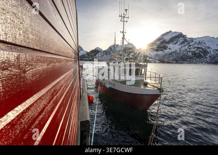 Barca da pesca nel porto con montagne innevate sullo sfondo, illuminate da raggi del sole, un peschereccio è ormeggiato su un molo di fronte a Snow-c Foto Stock