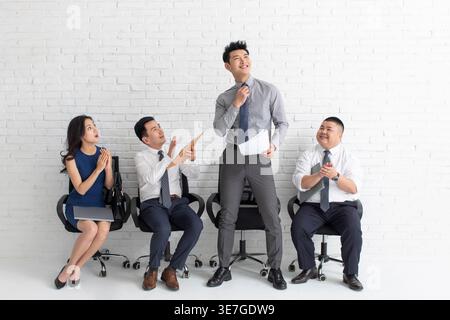 Sorridente candidato maschio cinese che tiene il suo curriculum e fissa la cravatta guardando lontano mentre viene applaudito da altri candidati di lavoro seduti contro il bianco B... Foto Stock