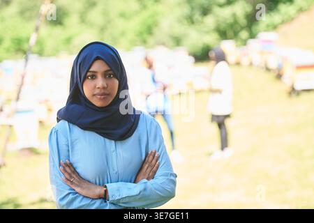 Donna musulmana seria che indossa hijab in piedi con le braccia incrociate nella fattoria di produzione del miele locale durante il giorno Foto Stock