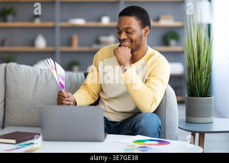 Web designer uomo nero appoggiato a mano sul mento guardando campioni di colore seduti sul divano davanti al tavolo con un computer portatile nel soggiorno di casa Foto Stock