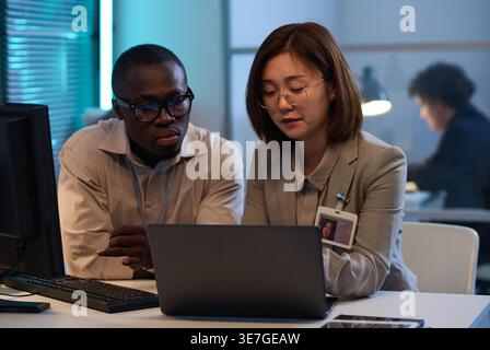 Programmatrici asiatiche e nere che si siedono alla scrivania discutendo di programmi per computer su un computer portatile che lavorano a tarda notte in ufficio Foto Stock