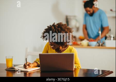 Donna nera sorridente con afro che scrive su un notebook seduto a un tavolo con un portatile e succo d'arancia vicino al marito in piedi al bancone della cucina a fette... Foto Stock