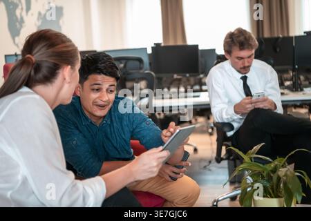 Uomo mediorientale che usa un tablet con una compagna di lavoro caucasica seduta vicino ai colleghi che usa un gadget nell'area relax dell'ufficio durante la pausa Foto Stock