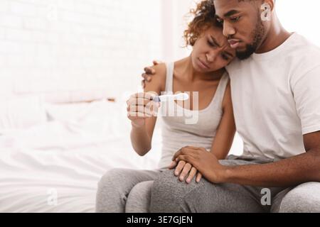 Coppia nera delusa che tiene un test di gravidanza negativo mentre si siede insieme a letto a casa Foto Stock