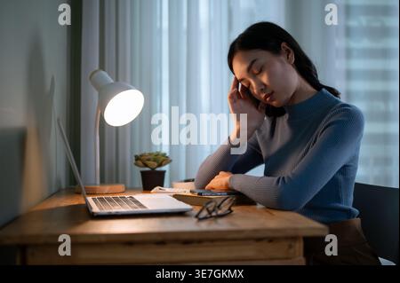 Donna asiatica seduta con gli occhi chiusi di fronte al laptop appoggiato faccia a mano stanca e assonnata nel salotto di casa Foto Stock