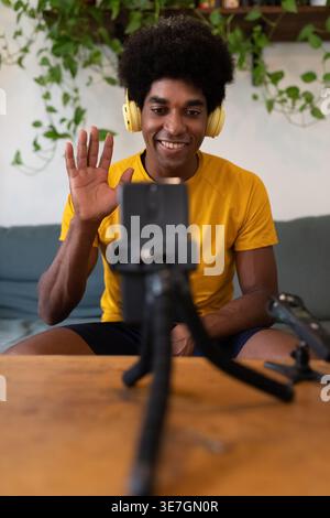 Smiling Black maschio influencer in cuffie vlogging utilizzando lo smartphone salutando i follower mentre si siede sul divano di casa Foto Stock