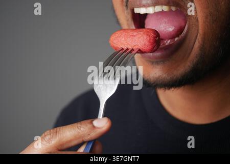 Primo piano uomo che mangia salsicce sulla forchetta mostrando bocca, lingua e mano Foto Stock