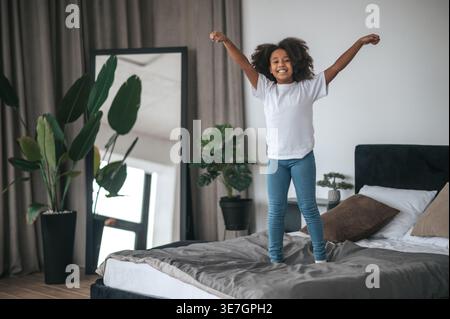 Allegra ragazza afroamericana con acconciatura afro che alza le mani mentre salta sul letto a casa Foto Stock