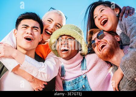 Allegra donna nera che indossa un cappello in piedi con uomini asiatici e caucasici che danno un giro in piggyback ad amici femminili che guardano all'aperto contro il cielo blu Foto Stock