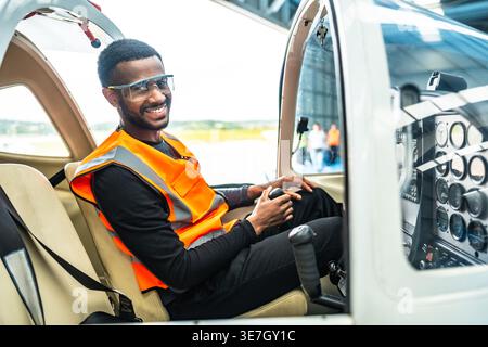 Un ingegnere afroamericano maschio sorridente che siede all'interno di un jet privato che tiene il controllo sulla cabina di pilotaggio nell'hangar Foto Stock