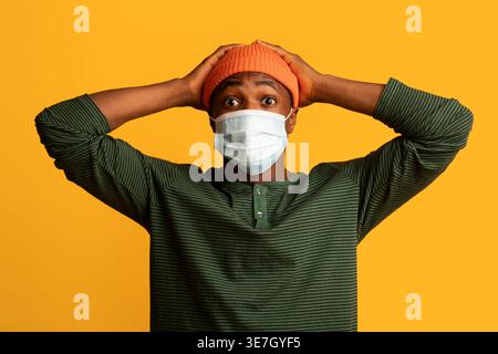 Scioccato uomo Nero che indossa berretto e maschera medica protettiva in piedi con le mani dietro la testa su sfondo giallo in studio Foto Stock