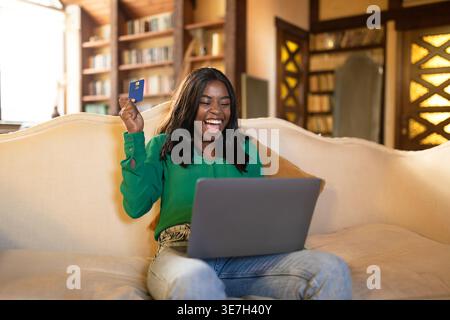 Allegra ragazza nera adolescente con laptop che regge la carta di credito urlando mentre si siede sul divano nel soggiorno di casa Foto Stock
