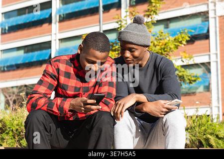 Amici maschi neri che si divertono a condividere lo smartphone mentre sono seduti sulla panchina in città nelle giornate di sole Foto Stock