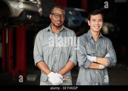 Meccanici maschi neri e asiatici che indossano guanti mentre sono in piedi insieme in officina di riparazione auto Foto Stock