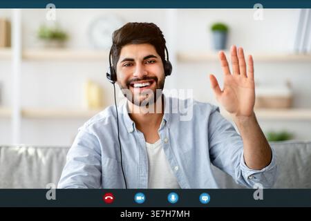Uomo arabo sorridente con la barba che indossa la cuffia che ondeggia la mano mentre videochiama sul laptop seduto sul divano nel soggiorno di casa Foto Stock