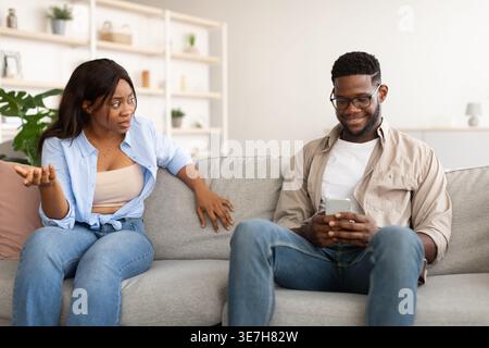 Infastidita la donna nera che guarda il marito sorridente usando lo smartphone seduto sul divano nel salotto di casa Foto Stock