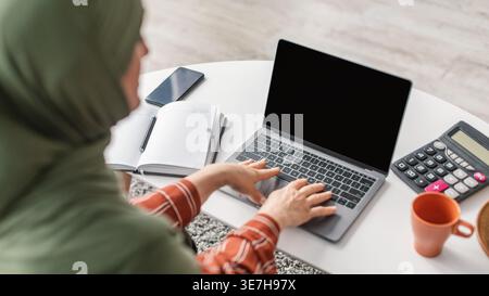 Immagine ritagliata di una donna musulmana mediorientale di mezza età in hijab che utilizza un computer portatile seduto al tavolo con una tazza da caffè, una calcolatrice, uno smartphone e ... Foto Stock