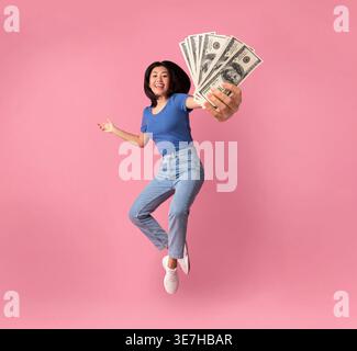 Donna asiatica sorridente che tiene e mostra banconote di carta mentre salta su sfondo rosa in studio Foto Stock