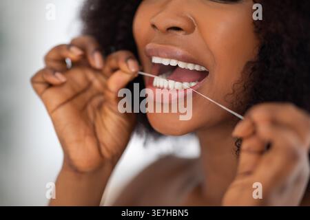Immagine ritagliata di donna nera con capelli ricci che le puliscono i denti usando il filo interdentale mentre sta in piedi a casa Foto Stock