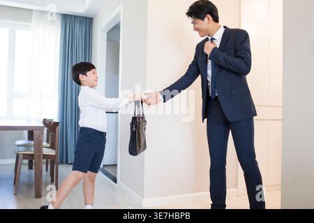 Un ragazzo cinese che dà al padre la sua borsa prima di partire per il lavoro in piedi a casa Foto Stock