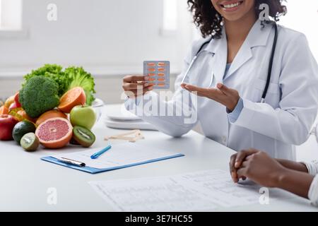 Immagine ritagliata di una nutrizionista nera con stetoscopio intorno al collo che raccomanda pillole al paziente seduto al tavolo con appunti, fr... Foto Stock