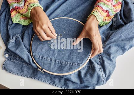 Sopra le mani angolate di artigiani di corse miste che fanno ricami con perline sul retro della giacca in denim in laboratorio Foto Stock
