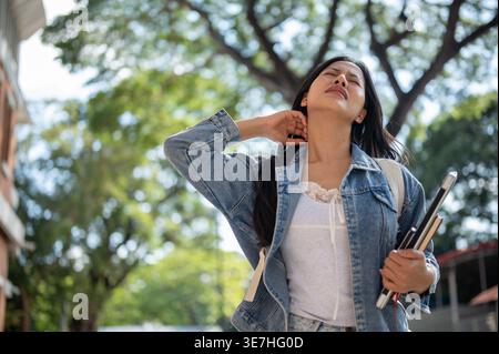 Studentessa universitaria thailandese stanca con gli occhi chiusi che indossa lo zaino che tiene il laptop e il libro che tocca il collo in piedi fuori dall'edificio universitario duri... Foto Stock