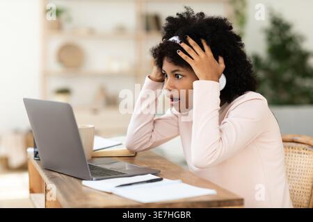 Ragazza preadolescente nera scioccata con afro che indossa le cuffie che guarda il laptop seduto alla scrivania in salotto Foto Stock