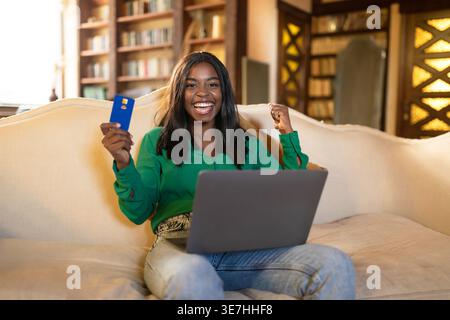 Adolescente nera con laptop in possesso della carta di credito che celebra la vittoria seduta sul divano nel soggiorno di casa Foto Stock
