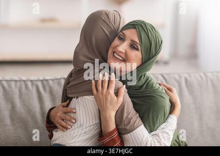 Allegra madre musulmana mediorientale di mezza età in hijab che abbraccia sua figlia mentre siede sul divano nel soggiorno di casa Foto Stock