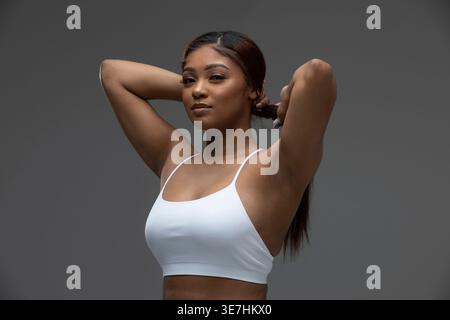 Donna nera che indossa una canotta bianca fissando i capelli su sfondo grigio in studio Foto Stock