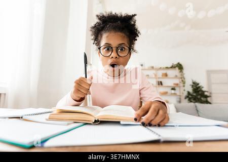 Scioccata ragazza nera preadolescente che indossa gli occhiali tenendo la penna seduta a tavola con un libro aperto a casa Foto Stock