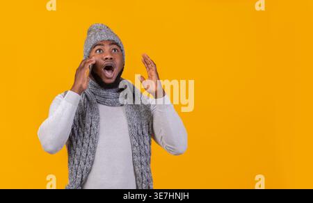 Scioccato uomo Nero che indossa berretto e sciarpa a maglia e che riceve una telefonata guardando lontano in piedi sullo sfondo giallo in studio Foto Stock