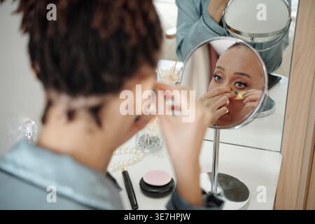 Donna nera che applica idratante sotto gli occhielli mentre è seduta a tavola davanti allo specchio a casa Foto Stock