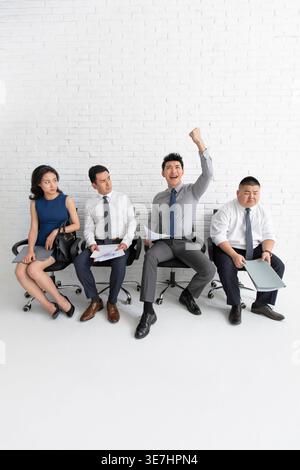 Allegro candidato cinese maschio che tiene il suo curriculum e solleva pugno chiuso mentre si siede insieme ad altri candidati di lavoro che lo guardano nel... Foto Stock